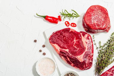 Set of various steaks with spices and herbs. Classic raw meat cuts includes ribeye, eye round and striploin steaks. Plaster white background, flat lay, top view