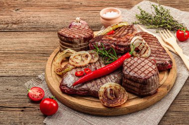 Grilled set of various steaks with vegetables, spices and herbs. Classic meat cuts includes ribeye, eye round, flank and striploin steaks. Wooden background, flat lay, copy space