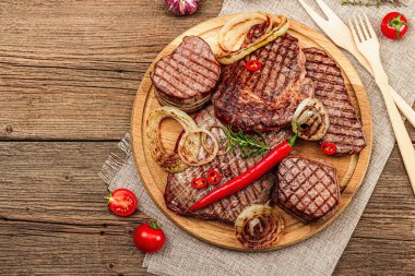 Grilled set of various steaks with vegetables, spices and herbs. Classic meat cuts includes ribeye, eye round, flank and striploin steaks. Wooden background, flat lay, top view