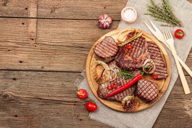 Grilled set of various steaks with vegetables, spices and herbs. Classic meat cuts includes ribeye, eye round, flank and striploin steaks. Wooden background, flat lay, top view