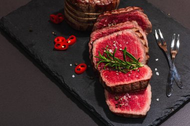 Rare cooking level Eye round steak with spices and herbs. Classic grilled meat cut ready to eat. Picnic or dinner concept. Hard light, dark shadow, black stone background, flat lay, close up