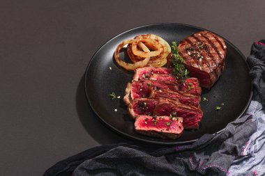 Rare cooking level Sirloin steak with spices and herbs. Classic grilled meat cut ready to eat. Picnic or dinner concept. Hard light, dark shadow, black stone background, flat lay, copy space