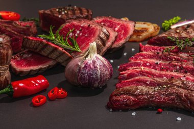 Grilled set of various steaks with vegetables, spices and herbs. Classic meat cuts includes ribeye, eye round, flank and striploin steaks. Black stone background, flat lay, close up