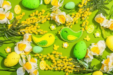 Easter composition with traditional spring flowers and handmade felt birds. Decorative eggs, rabbits, hard light, dark shadow, green background, top view