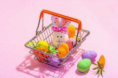 Easter sale concept. Shopping basket with festive symbols - rabbit, eggs, bird, and traditional decor. Pastel design, trendy hard light, dark shadow, pink background, copy space