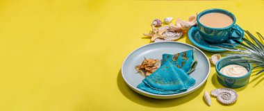 Deniz usulü bir fincan kahve ve ince mavi krep. Deniz yıldızı, deniz kabukları, palmiye yaprakları. Sert ışık, koyu gölge, parlak sarı arkaplan, afiş biçimi