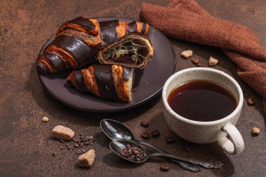 Bir fincan kahve ve koyu tonda çikolatalı kruvasan. Günaydın iyi ruh hali, Fransız kahvaltı konsepti. Esmer şeker, çatal bıçak, sert ışık, koyu gölge, taş beton arka plan, kapat