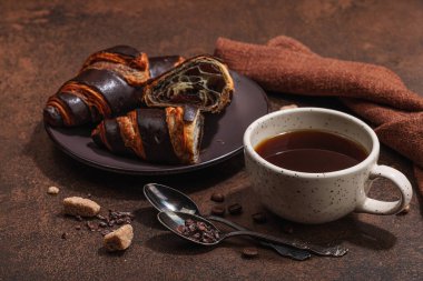 Bir fincan kahve ve koyu tonda çikolatalı kruvasan. Günaydın iyi ruh hali, Fransız kahvaltı konsepti. Esmer şeker, çatal bıçak, sert ışık, koyu gölge, taş beton arka plan, kapat