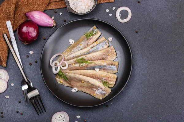 stock image Fillet of pickled marinated sea herring fish with shallots and dill. Cutlery, vintage napkin, hard light, dark shadow. Black stone concrete background, flat lay, top view