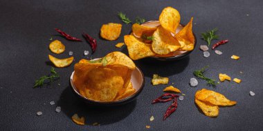 Kırmızı biber, maydanoz ve siyah beton arka planda tuz ile servis edilen ev yapımı patates cipsi kaseleri. Modaya uygun sert ışık, koyu gölge, afiş biçimi