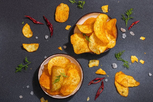 Kırmızı biber, maydanoz ve siyah beton arka planda tuz ile servis edilen ev yapımı patates cipsi kaseleri. Modaya uygun sert ışık, koyu gölge, üst manzara