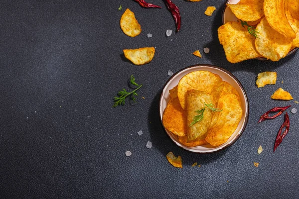 Kırmızı biber, maydanoz ve siyah beton arka planda tuz ile servis edilen ev yapımı patates cipsi kaseleri. Modaya uygun sert ışık, koyu gölge, üst manzara