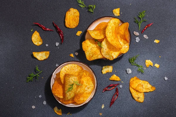 stock image Bowls of homemade potato chips served with chili pepper, parsley, seal salt on black stone concrete background. Trendy hard light, dark shadow, top view