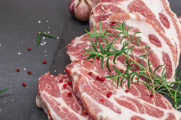 stock image Raw pork neck meat. Chop steak, red peppercorn, garlic cloves, sea salt and fresh rosemary. Trendy stand, black background, flat lay, close up