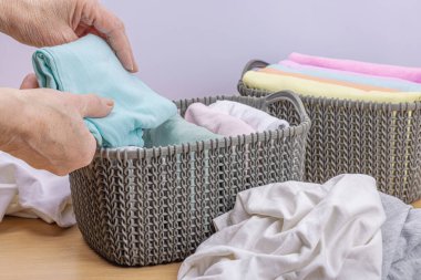 Kadınların elleri temiz giysileri katlar. Sepetlerde dikey giysi deposu var. Dolap temizliği, çevre dostu tarzda. Pastel renkli bebek tişörtleri. Hafif ahşap masa, boşluğu kopyala