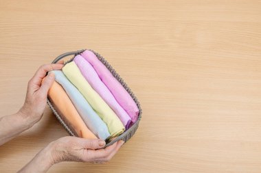 Kadınların elleri temiz giysileri katlar. Sepetlerde dikey giysi deposu var. Dolap temizliği, çevre dostu tarzda. Pastel renkli bebek tişörtleri. Hafif ahşap masa, üst manzara.