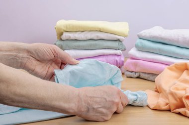 Kadınların elleri temiz giysileri katlar. Sepetlerde dikey giysi deposu var. Dolap temizliği, çevre dostu tarzda. Pastel renkli bebek tişörtleri. Hafif ahşap masa, boşluğu kopyala