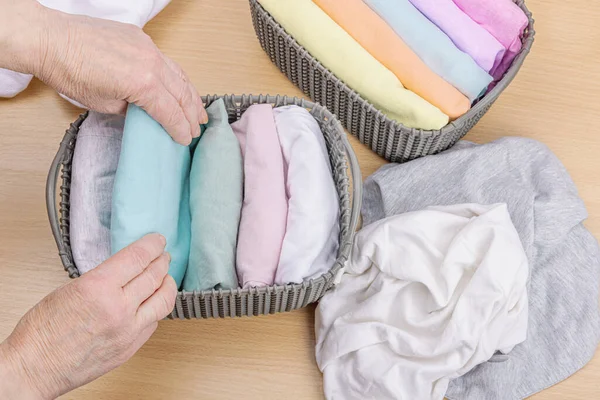 Stock image Women's hands fold clean clothes. Vertical storage of clothing in the baskets. Closet cleaning, eco friendly style. Pastel colored baby T-shirts. Light wooden table, top view