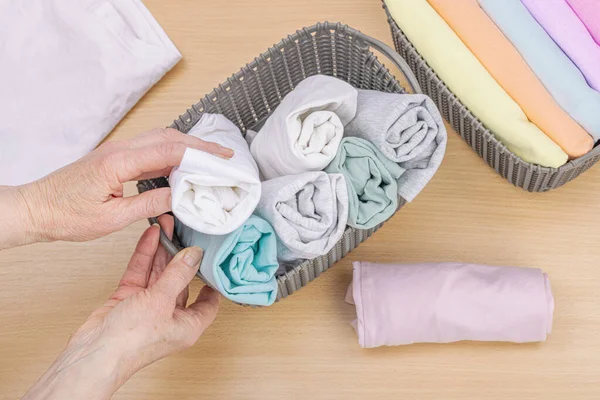 Stock image Women's hands fold clean clothes. Vertical storage of clothing in the baskets. Closet cleaning, eco friendly style. Pastel colored baby T-shirts. Light wooden table, top view