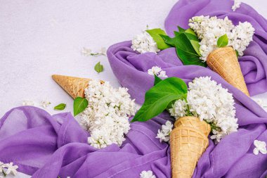 Beyaz leylak çiçekleri kremalı dondurma külahları içinde beton zeminde. Düz yatak, mor peçete, geleneksel bahar konsepti, kopyalama alanı