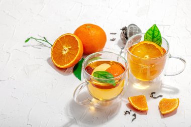Cam bardakta portakal çayı ve olgun meyveler. Serinletici mevsimlik içecek, yeşil narenciye yapraklı kavramsal arka plan. Modern biçim, seçici odak, sert ışık, karanlık gölge, kopyalama alanı