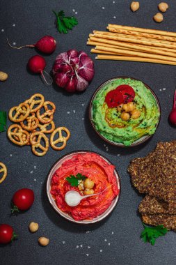 Çeşitli humus sosları, taze sebzeler ve otlar. Çıtır ekmekler, modern vegan sağlıklı yiyecekler. Sert ışık, karanlık gölge, siyah taş arka plan, üst görüş