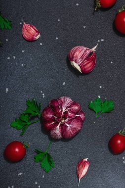Taze sebzeler ve otlarla mutfak arka planı. Domates, kiraz, sarımsak, deniz tuzu ve maydanoz. Düz yatıyordu, sert ışık, karanlık gölge, siyah taş beton zemin