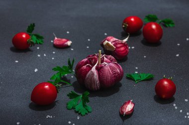 Taze sebzeler ve otlarla mutfak arka planı. Domates, kiraz, sarımsak, deniz tuzu ve maydanoz. Düz yatıyordu, sert ışık, karanlık gölge, siyah taş beton zemin