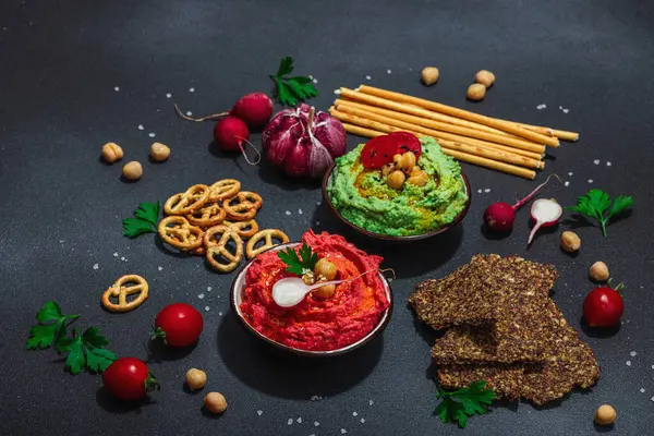 Stock image Various hummus dips, fresh vegetables and herbs. Crispy bread snacks, modern vegan healthy food. Hard light, dark shadow, black stone concrete background, copy space