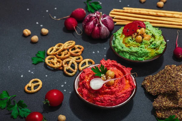 stock image Various hummus dips, fresh vegetables and herbs. Crispy bread snacks, modern vegan healthy food. Hard light, dark shadow, black stone concrete background, close up