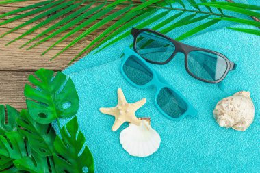 Yaz tatili kavramı. Banyo havlusu, güneş gözlüğü, deniz dekoru. Palmiye ve canavar yaprakları, ahşap zemin, düz yatış, üst manzara