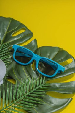 Yaz tatili kavramı. Güneş gözlüğü, deniz dekoru, palmiye ve canavar yaprakları, orkide çiçeği. Parlak sarı arkaplan, sert ışık, koyu gölge, düz uzanma, kopyalama alanı