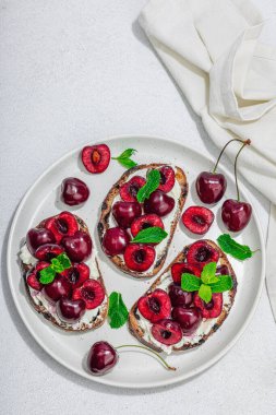 Tatlı kirazlı, krem peynirli ve nane yapraklı taze ekmek sandviçleri. Günaydın kahvaltı konsepti. Sert ışık, karanlık gölge, beyaz taş arka plan, üst görüş