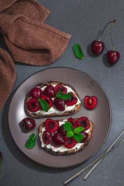 Tatlı kirazlı, krem peynirli ve nane yapraklı taze ekmek sandviçleri. Günaydın kahvaltı konsepti. Sert ışık, karanlık gölge, siyah taş arka plan, üst görüş