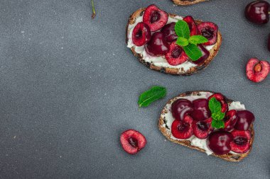 Tatlı kirazlı, krem peynirli ve nane yapraklı taze ekmek sandviçleri. Günaydın kahvaltı konsepti. Sert ışık, karanlık gölge, siyah taş arka plan, üst görüş