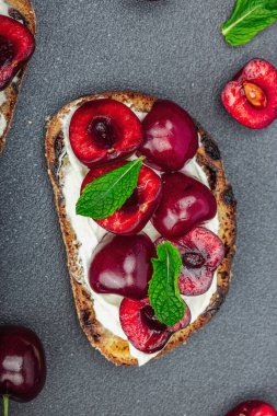 Tatlı kirazlı, krem peynirli ve nane yapraklı taze ekmek sandviçleri. Günaydın kahvaltı konsepti. Sert ışık, karanlık gölge, siyah taş arka plan, üst görüş