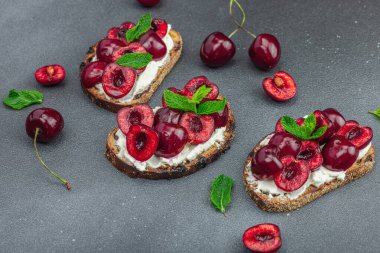 Tatlı kirazlı, krem peynirli ve nane yapraklı taze ekmek sandviçleri. Günaydın kahvaltı konsepti. Sert ışık, karanlık gölge, siyah taş zemin, kopyalama alanı