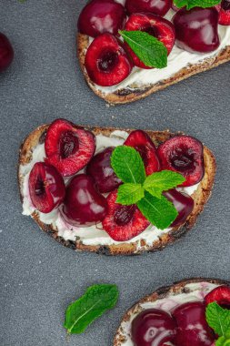 Tatlı kirazlı, krem peynirli ve nane yapraklı taze ekmek sandviçleri. Günaydın kahvaltı konsepti. Sert ışık, karanlık gölge, siyah taş arka plan, üst görüş