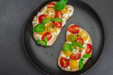 Domates kirazlı, krem peynirli ve fesleğenli taze ekmek sandviçleri. Sabah kahvaltısı kavramı. Sert ışık, karanlık gölge, siyah taş arka plan, üst görüş