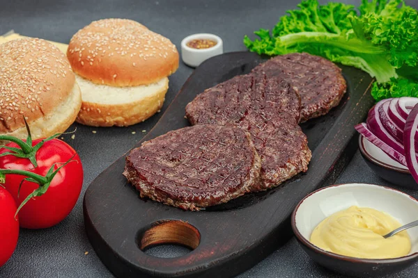 Grilled burger patties. Hot beef cutlets, buns, vegetables and sauce. Homemade American classic, traditional food for picnic, party or Independence Day. Dark stone concrete background, close up