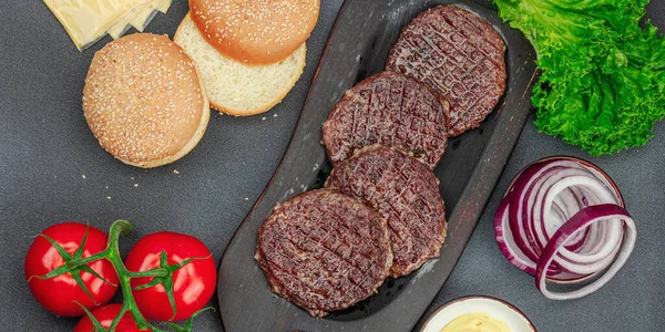 Grilled burger patties. Hot beef cutlets, buns, vegetables and sauce. Homemade American classic, traditional food for picnic, party or Independence Day. Dark stone concrete background, banner format