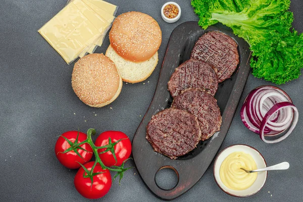 Grilled burger patties. Hot beef cutlets, buns, vegetables and sauce. Homemade American classic, traditional food for picnic, party or Independence Day. Dark stone concrete background, top view