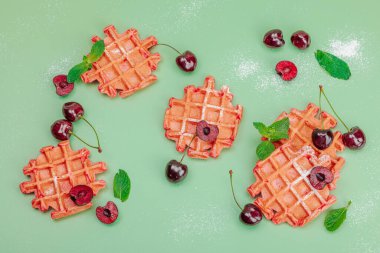 Homemade Belgian Waffles with sweet cherries. Gluten-free red dessert, fresh fruits and basil. Sweet sugar powder, trendy green background, top view