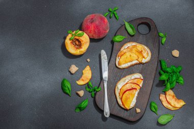 Tatlı şeftali dilimleri, krem peynir ve fesleğen yapraklı taze ekmek sandviçleri. Günaydın kahvaltı konsepti. Sert ışık, karanlık gölge, siyah taş arka plan, üst görüş