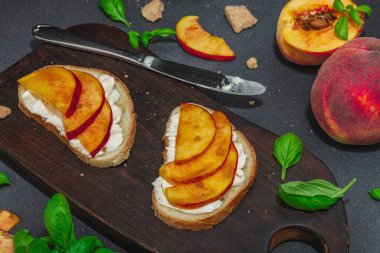 Tatlı şeftali dilimleri, krem peynir ve fesleğen yapraklı taze ekmek sandviçleri. Günaydın kahvaltı konsepti. Sert ışık, karanlık gölge, siyah taş beton arka plan, kapat