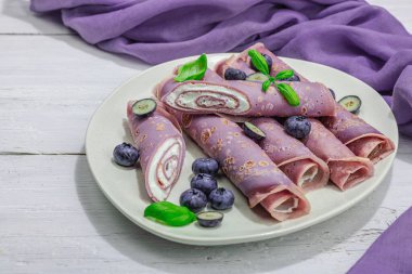 Kremalı ve yaban mersinli ince mor krepler. Sağlıklı vegan, laktoz ve glütensiz yemek, kahvaltı konsepti. Modaya uygun sert ışık, karanlık gölge, beyaz ahşap arka plan, kapat