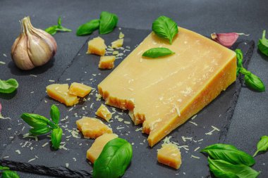 Modern tezgahta bir parça parmesan peyniri. Sarımsak, taze fesleğen, sağlıklı yemek için geleneksel İtalyan malzemeleri. Siyah taş zemin, düz zemin, yakın plan.