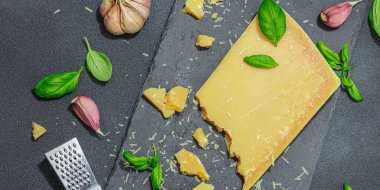 Modern tezgahta bir parça parmesan peyniri. Sarımsak, taze fesleğen, sağlıklı yemek için geleneksel İtalyan malzemeleri. Siyah taş zemin, düz zemin, pankart biçimi