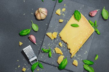 Modern tezgahta bir parça parmesan peyniri. Sarımsak, taze fesleğen, sağlıklı yemek için geleneksel İtalyan malzemeleri. Siyah taş zemin, düz zemin, üst manzara
