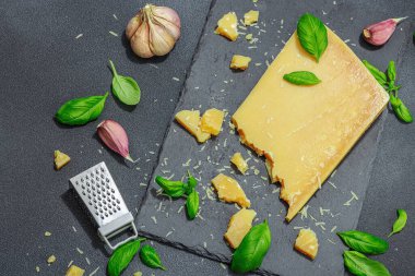 Modern tezgahta bir parça parmesan peyniri. Sarımsak, taze fesleğen, sağlıklı yemek için geleneksel İtalyan malzemeleri. Siyah taş zemin, düz zemin, üst manzara